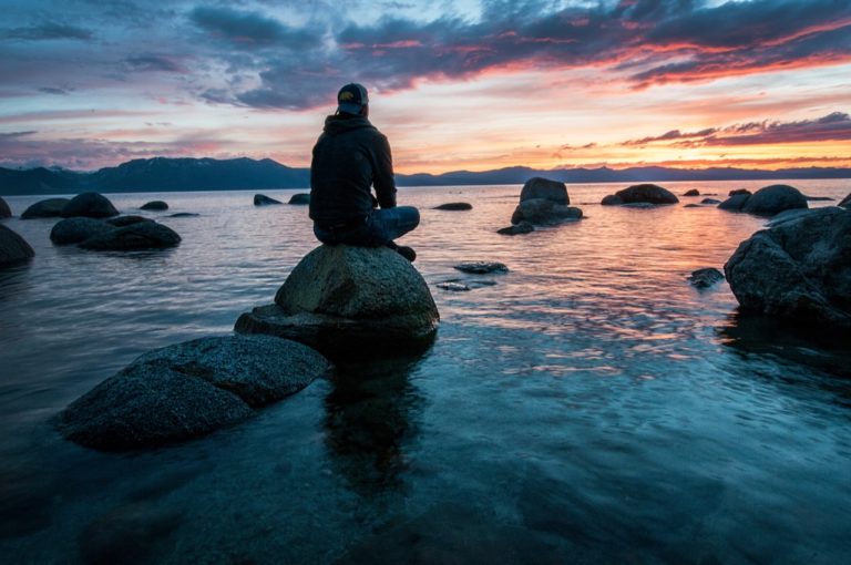 Person sitting on a rock out in the water looking at the sunset How to thrive as an empath